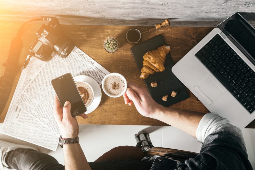 An aerial view of a messy desk with a laptop. A man is seated at it drinking a coffee and looking at his cellphone.